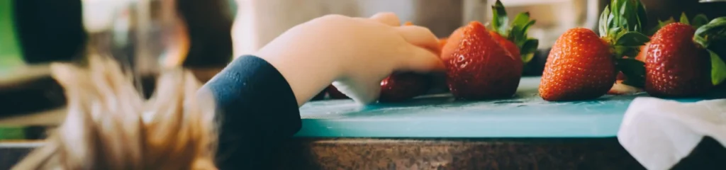 An image of a child reaching over the kitchen countertop to grab some strawberries.